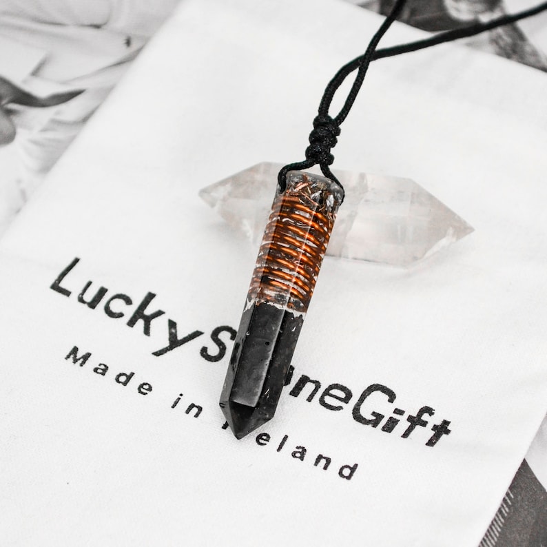May include: Black and copper crystal pendant necklace with a black cord. The pendant is a pointed crystal with copper wire wrapped around the center. The necklace is on a white surface with the text "LuckyStoneGift Made in Ireland".