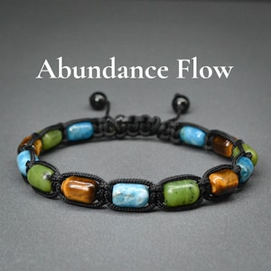 May include: A black cord bracelet with colorful, rectangular beads. The beads are in shades of blue, green, and brown. The text "Abundance Flow" is displayed in white above the bracelet.