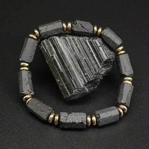 Puede incluir: Pulsera de turmalina negra con detalles dorados. La pulsera está hecha de cuentas de turmalina negra y separadores dorados. La pulsera se muestra sobre un fondo negro con un gran cristal de turmalina negra.