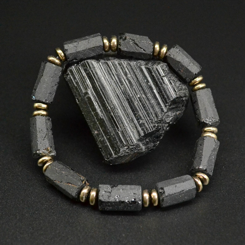May include: Black Tourmaline bracelet with gold accents. The bracelet is made of black tourmaline beads and gold spacers. The bracelet is shown on a black background with a large black tourmaline crystal.