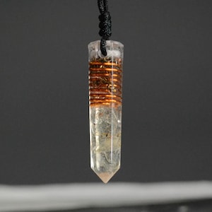 Puede incluir: Un colgante de cristal de cuarzo transparente con un alambre de cobre envuelto alrededor. El colgante está unido a un cordón negro.