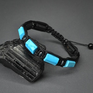 Peut inclure: Un bracelet macramé noir orné de pierres rectangulaires de couleur turquoise. Le bracelet est présenté sur une surface sombre et texturée. Il possède une fermeture coulissante pour un ajustement facile.