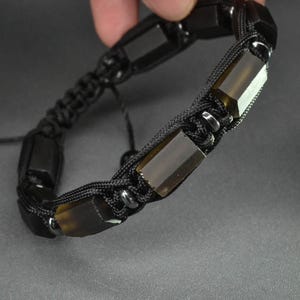Puede incluir: Pulsera trenzada negra con piedras rectangulares de color oscuro. La pulsera tiene un diseño de macramé con nudos corredizos ajustables. Las piedras son translúcidas, sobre un fondo gris.