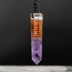 Puede incluir: Un colgante de cristal de amatista púrpura con un alambre de cobre envuelto alrededor del centro, colgando de un cordón negro.