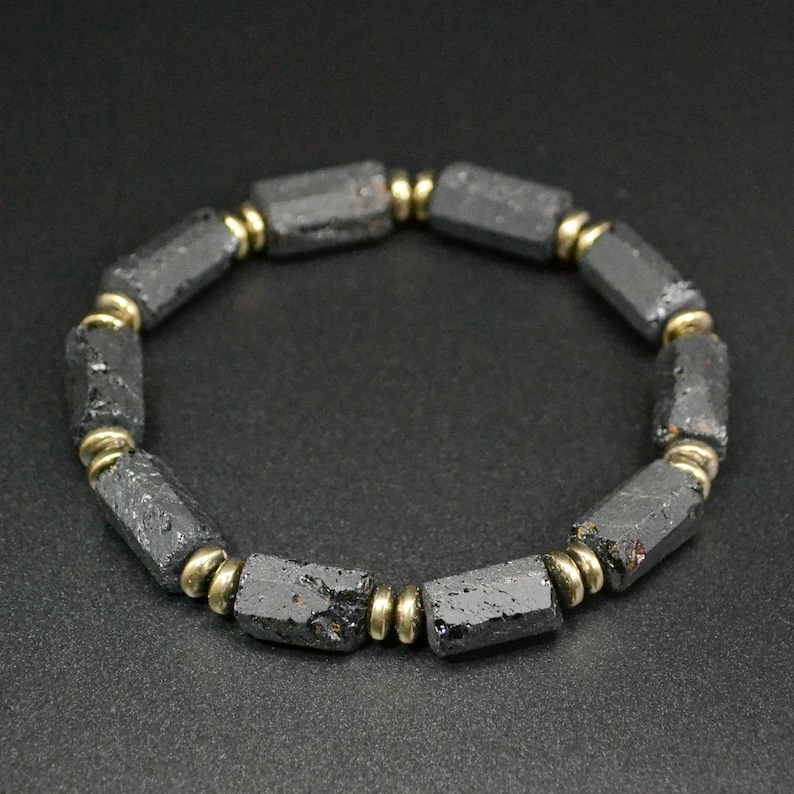 May include: A black beaded bracelet with gold accents. The beads are made of black lava rock and are shaped like small cylinders. The bracelet is made of natural materials and has a rustic look.