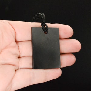 Puede incluir: Un colgante rectangular negro con un agujero en la parte superior, colgado de un cordón negro.