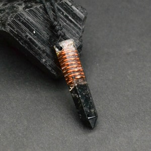 May include: Black crystal point pendant with a copper wire wrapped around it, set in clear resin. The pendant is on a black cord and is hanging from a black rock.