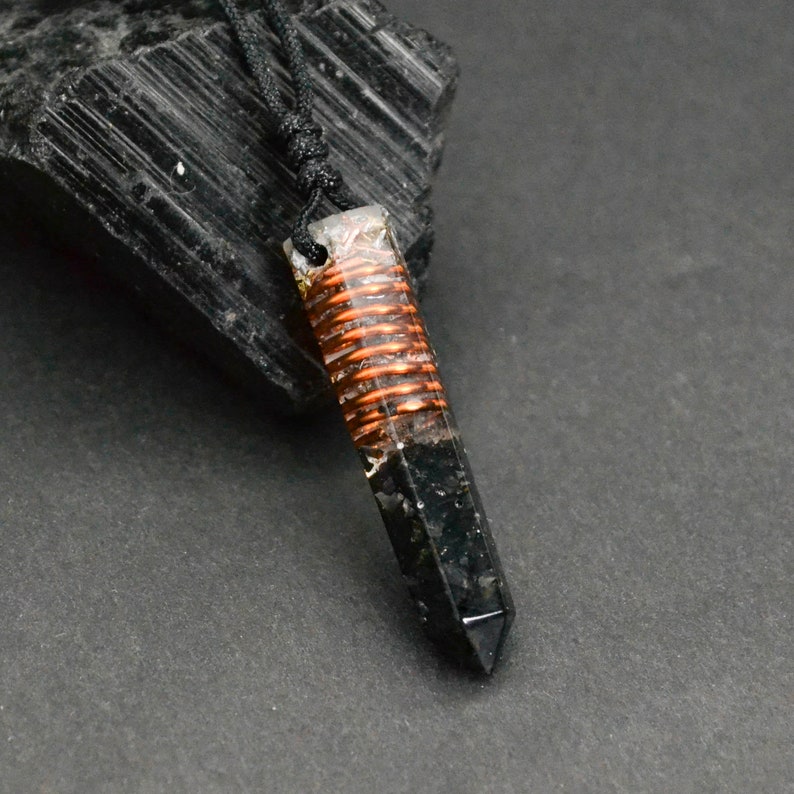 May include: Black crystal point pendant with a copper wire wrapped around it, set in clear resin. The pendant is on a black cord and is hanging from a black rock.