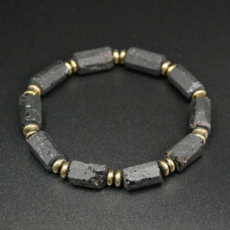 May include: A black beaded bracelet with gold accents. The beads are made of black lava rock and are shaped like small cylinders. The bracelet is made of natural materials and has a rustic look.