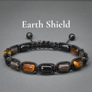 Puede incluir: Pulsera de macramé negra con cuentas rectangulares negras, marrones y color ámbar. La pulsera es ajustable con dos cuentas negras en los extremos de los cordones. El texto "Earth Shield" se muestra en blanco.