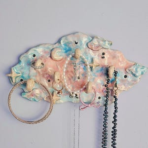 Op de afbeelding: Een wolkvormige keramische wandorganizer met een gemarmerd roze en blauw glazuur, voorzien van kleine uitstekende haakjes en decoratieve ster- en maanaccenten. Het houdt diverse sieraden vast, waaronder een gouden armband, kettingen en ringen.