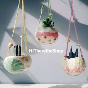 May include: Three hanging planters with colourful designs and plants. The planters are made of ceramic and have a unique, hand-painted style. The plants are a cactus, a succulent, and a green plant.