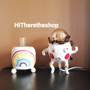 Puede incluir: Dos lámparas de cerámica, una con forma de arcoíris con una bombilla en su interior y la otra con forma de personaje de dibujos animados con una bombilla en su interior. Ambas lámparas son blancas con detalles coloridos.