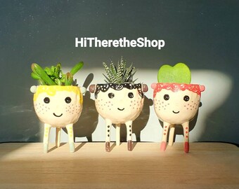 May include: Three ceramic planters shaped like faces with painted details. Each planter has a different colored face and a different type of plant growing inside. The planters are sitting on a wooden surface.
