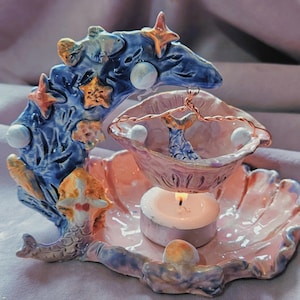 May include: A ceramic wax warmer featuring a blue crescent moon base adorned with gold starfish and a mermaid tail. A pink shell-shaped dish hangs from the moon by a copper wire, positioned above a small white tea light candle on a pink base.