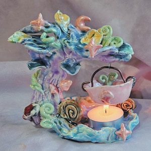 May include: A ceramic candle holder with a whimsical, undersea theme. The holder features a coral-like structure in shades of blue, green, and purple, adorned with starfish and other sea-inspired elements. A small pink bowl hangs from the top, and a lit candle sits below.