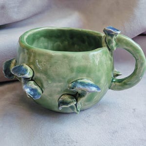Op de afbeelding: Een groene keramische mok met een handvat en blauwe paddenstoeldetails. De mok is handgemaakt en heeft een rustieke, aardse uitstraling.