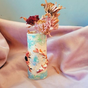 Puede incluir: Un jarrón de cerámica blanca con un patrón floral azul y verde y peces koi pintados. El jarrón está lleno de flores secas.