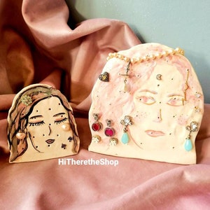 Puede incluir: Dos soportes de exhibición de aretes de cerámica con forma de cara con detalles pintados. Los soportes tienen múltiples agujeros para exhibir aretes. Los soportes están sobre una superficie rosa.