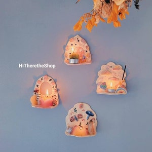 May include: Four ceramic candle holders with whimsical designs are mounted on a light blue wall. Each holder features a lit candle, and the designs include a cactus, mushrooms, and a rainbow. The shop name "HiTheretheShop" is visible.
