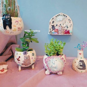 Könnte beinhalten: Fünf Keramik-Blumentöpfe mit Tier- und Blumenmotiven. Die Töpfe sind in verschiedenen Formen und Größen, darunter ein Hängetöpf, eine Vase und ein Topf mit Beinen. Die Töpfe sind mit handgemalten Details verziert, darunter Bären, Katzen und Blumen.