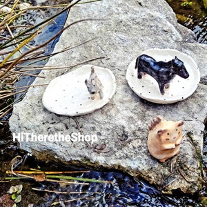 May include: Three ceramic animal figurines on white dishes with black polka dots. A black cow, a gray dog, and a brown cat.