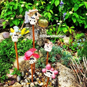 Puede incluir: Cinco estacas de jardín de cerámica en forma de hongo con caras pintadas y detalles coloridos. Los hongos están en estacas de madera marrón y están dispuestos en un jardín con rocas y plantas verdes.