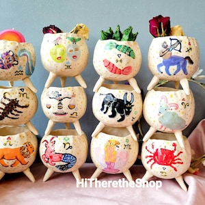 May include: A set of 12 ceramic planters, each featuring a different zodiac sign. The planters are painted in bright colors and have a whimsical, hand-painted style. The planters are stacked in a pyramid shape, with the top planter being a Sagittarius.