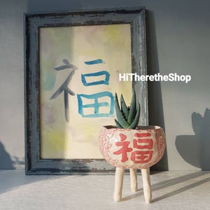 May include: A white ceramic planter with three legs and a red and white design featuring the Chinese character for "good fortune" or "happiness". The planter is filled with a small succulent plant.