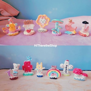 May include: A collection of 10 hand-painted ceramic figurines featuring whimsical characters like a bear, a bunny, a rainbow, and a cactus. The figurines are painted in bright colors and have a cute, cartoonish style.