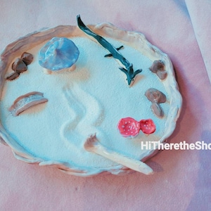 Puede incluir: Una bandeja de cerámica blanca con una superficie similar a la arena, que presenta un hongo azul, un pequeño puente, un hueso blanco, dos hongos rojos y una figurita de lagarto negro y verde. La bandeja está rodeada por un borde texturizado.