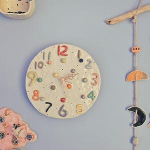 Puede incluir: Colección de decoraciones de pared de cerámica caprichosas sobre una pared azul claro. Los artículos incluyen un reloj redondo con números coloridos, un móvil con formas de estrella y luna y otras piezas decorativas. El reloj mide aproximadamente 20 cm de diámetro.