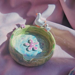 May include: A ceramic bowl with a light green rim and a blue and white interior. The bowl is decorated with a pink flower and small white duck figurines. The bowl is on a pink surface.