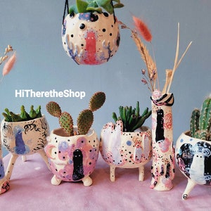 May include: A collection of five hand-painted ceramic planters with various plants, including succulents and cacti. The planters feature whimsical designs with colourful details and three-dimensional legs. The text "HiThereTheShop" is visible in the image.