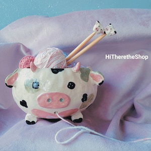 Puede incluir: Un bol de lana de cerámica con forma de vaca, con manchas blancas y negras, orejas rosas y una nariz rosa. El bol está lleno de lana rosa y blanca y dos agujas de tejer de madera con tapas en forma de vaca. El bol está sentado sobre una tela de color púrpura claro.