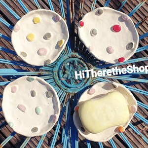 May include: Four white ceramic soap dishes with colorful dots. The dishes are arranged in a circle on a blue metal surface. One dish has a bar of soap inside.