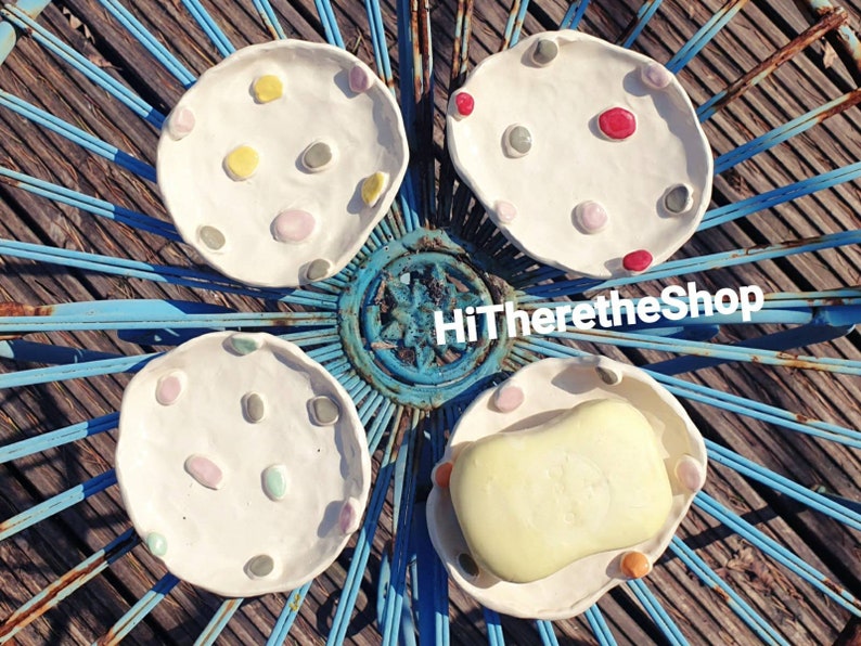 May include: Four white ceramic soap dishes with colorful dots. The dishes are arranged in a circle on a blue metal surface. One dish has a bar of soap inside.
