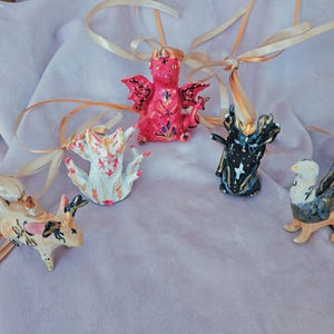 Puede incluir: Cinco figuritas de cerámica con detalles intrincados y acentos dorados. Las figuritas son un dragón rojo, un gato blanco, un pájaro negro y dorado, una criatura negra y dorada con alas y un conejo marrón. Cada figurita está unida a una cinta dorada.