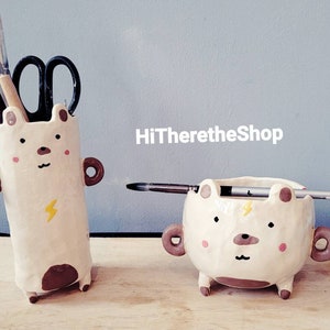 Puede incluir: Dos porta lápices de cerámica con forma de oso. El porta lápices más grande está de pie y tiene un diseño de rayo amarillo. El porta lápices más pequeño está acostado y tiene un pincel apoyado encima. Ambos porta lápices tienen detalles marrones y caras lindas.