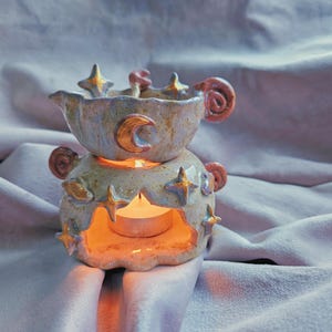 Puede incluir: Un calentador de cera de cerámica con diseño de dos piezas y esmalte moteado en azul claro y crema. El exterior está decorado con estrellas y lunas crecientes pintadas en oro en relieve, con una vela de té encendida visible en la base.