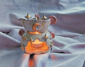 May include: A ceramic wax warmer featuring a two-piece design with a speckled light blue and cream glaze. The exterior is decorated with raised gold-painted stars and crescent moons, with a lit tea light candle visible inside the base.