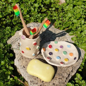 May include: A white ceramic toothbrush holder with colorful polka dots, three rainbow-colored toothbrushes, and a yellow bar of soap on a stone surface.