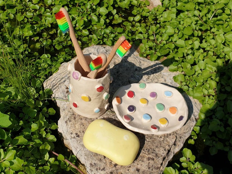 May include: A white ceramic toothbrush holder with colorful polka dots, three rainbow-colored toothbrushes, and a yellow bar of soap on a stone surface.
