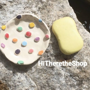 May include: A white ceramic dish with colorful dots and a yellow soap bar. The dish is on a gray rock surface. HiThereTheShop is written in white text on the rock.