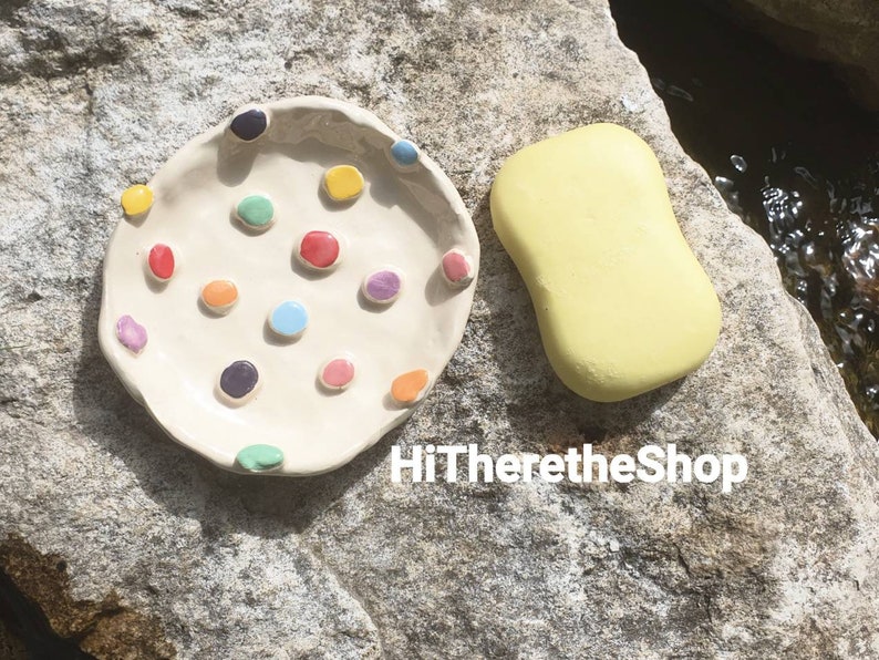 May include: A white ceramic dish with colorful dots and a yellow soap bar. The dish is on a gray rock surface. HiThereTheShop is written in white text on the rock.