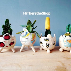 May include: Four whimsical, hand-painted ceramic planters, each with a unique design and small legs. One features ladybugs, another a unicorn, a third has bees, and the last a monkey. The planters hold various succulents and cacti.