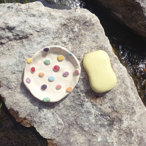 May include: A white ceramic dish with colorful polka dots, perfect for holding soap. The dish is sitting on a gray rock next to a yellow bar of soap.