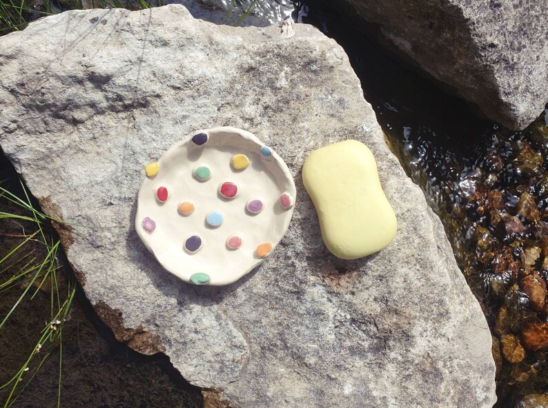 May include: A white ceramic dish with colorful polka dots, perfect for holding soap. The dish is sitting on a gray rock next to a yellow bar of soap.