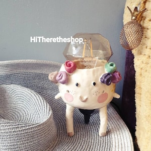 Puede incluir: Una lámpara de cerámica blanca con forma de criatura sonriente con mejillas rosadas y una bombilla en forma de diamante. La criatura tiene flores moradas, verdes y rosas en la cabeza. La lámpara está sentada sobre un sombrero de paja gris.