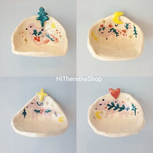 Puede incluir: Conjunto de cuatro platos de cerámica para joyas con diseños de animales caprichosos. Cada plato presenta un animal diferente con un diseño único, incluyendo un árbol, una luna creciente, una estrella y un corazón. Los platos están pintados en una suave combinación de rosa y blanco con acentos verdes, amarillos y rojos.